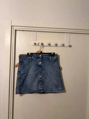 Blå jeanskjol med knappar framtill - Säljer en klassisk blå jeanskjol med knappar hela vägen framtill och bälteshällor i midjan. Kjolen har en kort passform och är gjord i denim, perfekt för en avslappnad och trendig look.