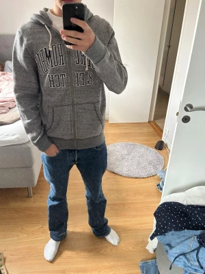 Grå hoodie från Abercrombie & Fitch - Snygg grå zip-hoodie från Abercrombie & Fitch med stor logga på bröstet. Hoodien har dragsko i huvan, två fickor framtill och ribbade muddar vid ärmar och nederkant. Perfekt för en avslappnad och trendig look.
