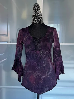 Lila boho-blus från KappAhl - Supersnygg lila blus från KappAhl med boho-vibbar, spetsdetaljer och snörning framtill. Blusen har utsvängda långärmar och ett unikt mönster i olika lila nyanser. Perfekt för dig som gillar en lite mer alternativ stil.