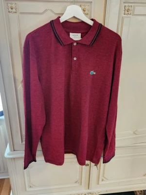 Vinröd långärmadpiké från Lacoste XXL Slimfit  - Vinröd långärmadpikétröja från Lacoste med klassisk krage och två knappar framtill. Tröjan har det ikoniska gröna krokodilmärket broderat på bröstet och svarta detaljer på kragen. Perfekt för en stilren och avslappnad look.
