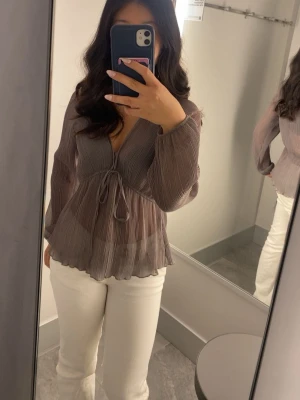 Blus från Zara - Snygg  blus i taupe från Zara med v-ringning och knytning framtill. Blusen har långa, luftiga ärmar och är tillverkad i ett tunt, transparent material som ger en lätt och flowy känsla. Perfekt att styla med jeans eller kjol.