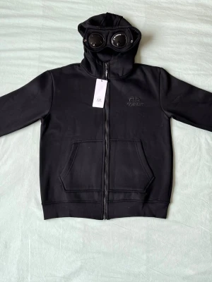 Svart hoodie med goggles från C.P. Company - Säljer en svart hoodie från C.P. Company med dragkedja och ikoniska goggles i huvan. Hoodien har känguruficka framtill och broderad logga på bröstet. Perfekt för dig som vill sticka ut med en unik streetwear-look.