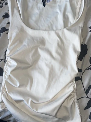 Vit baddräkt med rynkade sidor - Snygg vit baddräkt med smala axelband och rynkade detaljer längs sidorna. Klassisk rundad urringning fram och bak. Tillverkad i ett stretchigt, glansigt material som ger en bekväm passform och modern look. 