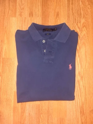 Polo Ralph Lauren Piké Långärmad - Klassisk mörkblå långärmad pikétröja från Polo Ralph Lauren med två knappar och rosa broderad logga på bröstet. Tillverkad i mjuk bomull och har en stilren krage. Perfekt för dig som gillar en enkel men snygg look. Finns inga defekter.