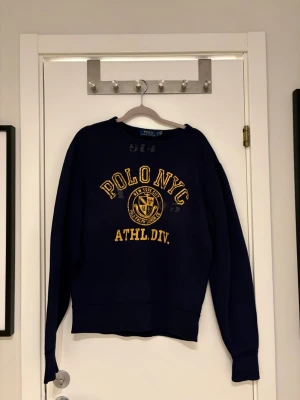 Mörkblå sweatshirt Polo NYC - Mörkblå sweatshirt från Polo Ralph Lauren med gul text 'POLO NYC ATHL. DIV.' och emblem framtill. Klassisk crewneck-modell med långa ärmar och ribbade muddar. Perfekt för en sportig och avslappnad stil. Strorlek s| skick 9/10| Pm för mer info😀