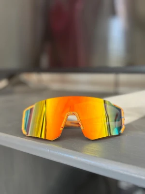 Barnstorlek Orange sportiga solglasögon - Säljer ett par sportiga solglasögon med spegelglas i orange och gult. Bågarna är också orange med stänk av andra färger. Glasögonen har en futuristisk, kantig form och täcker mycket av ansiktet – perfekta för en aktiv livsstil.