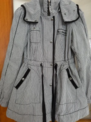  vindjacka - Snygg vindjacka med svartvita smala ränder, dragkedja och knappar . Jackan har flera fickor samt  Innerficka på vänster sida. Materialet känns lätt och passar perfekt för blåsiga dagar.