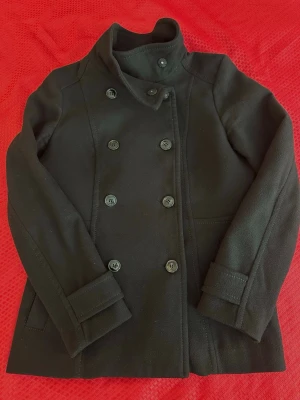 Svart dubbelknäppt trenchcoat HM st. 40 - Stilren svart trenchcoat med dubbelknäppning och hög krage. Jackan har raka fickor och dekorativa detaljer vid ärmsluten. Tillverkad i ett tjockare tyg som ger en klassisk look. Perfekt för dig som gillar tidlös och enkel stil.
