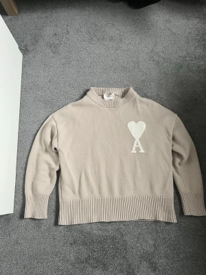 Beige stickad tröja från Ami Paris - Snygg beige stickad tröja från Ami Paris med ikoniskt vitt hjärta och bokstaven A på bröstet. Tröjan har ribbade muddar och är gjord i mjukt material. Perfekt för dig som gillar stilrena och trendiga plagg.