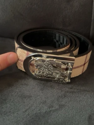 Burberry bälte  - Snyggt beige bälte från Burberry med klassiskt rutigt mönster i brunt och rött. Bältet har ett detaljerat metallspänne med Burberrys ikoniska riddare. Perfekt accessoar för att lyfta din outfit.