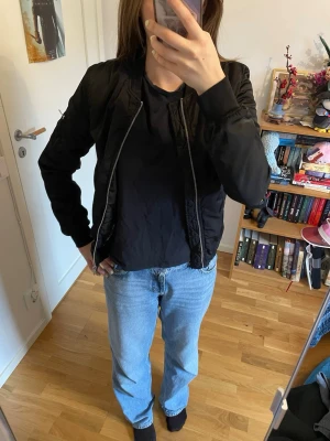 Svart bomberjacka från Lindex - Snygg svart bomberjacka från Lindex med blommigt foder i rosa och orange toner. Jackan har dragkedja framtill, ribbade muddar vid ärmar och midja samt en ficka med dragkedja på ena ärmen. Perfekt för dig som vill ha en stilren och trendig look.