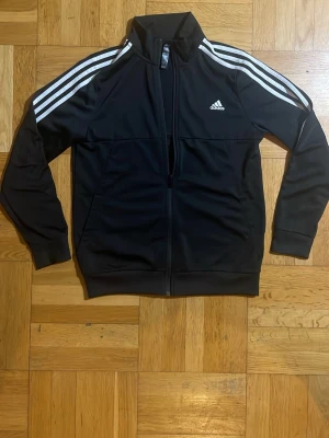 Svart Adidas track jacket med vita ränder - Svart track jacket från Adidas med klassiska vita ränder längs ärmarna och vit Adidas-logga på bröstet. Jackan har dragkedja framtill, hög krage och muddar vid ärmslut och midja. Perfekt för träning eller chill. Materialet är troligtvis polyester.