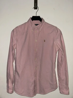 Rosa skjorta från Polo Ralph Lauren - Klassisk rosa skjorta från Polo Ralph Lauren i bomull med button-down krage och broderad logga på bröstet. Skjortan har lång ärm, custom fit och knappar framtill. Perfekt för en clean och stilren look. Pris kan diskuteras 