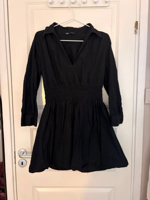 Svart kort klänning från Zara - Snygg svart kort klänning från Zara med v-ringning, markerad midja med smock och krage. Klänningen har långa ärmar och lätt utställd kjol. Perfekt för dig som gillar enkel men stilren design.