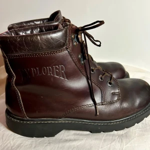 Mörkbruna läderkängor Explorer - Säljer ett par mörkbruna kängor från Explorer i äkta läder. Kängorna har kraftig sula, snörning med metallhål och vadderad kant upptill för extra komfort. Klassisk outdoor-look med Explorer-logga präglad på sidan. Perfekta för dig som gillar robust stil.