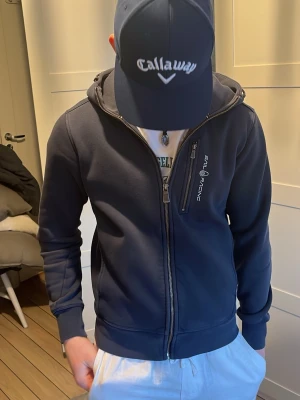 Mörkblå hoodie från Sail Racing - Snygg mörkblå hoodie från Sail Racing med dragkedja framtill och en extra bröstficka med zip. Hoodien har en stilren design, ribbade muddar och en skön huva. Perfekt för dig som gillar sportig och clean stil.