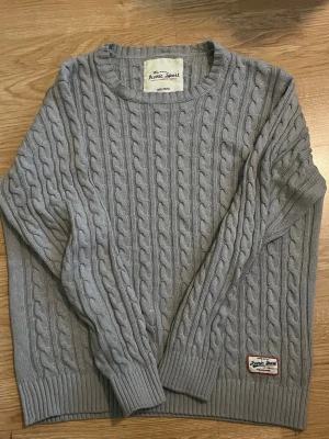 Grå kabelstickad tröja Iconic Sport - Snygg grå kabelstickad tröja från Iconic Sport. Tröjan har rund halsringning, ribbade muddar och en patch med logga nertill. Perfekt nu med våren runt hörnet. Strl S men passar mer som M