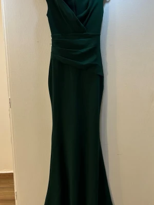 Mörkgrön långklänning från Goddiva - Elegant mörkgrön långklänning från Goddiva London med omlottdesign och markerad midja. Klänningen har en djup v-ringning och är ärmlös, vilket ger en stilren och sofistikerad look. Perfekt för speciella tillfällen.