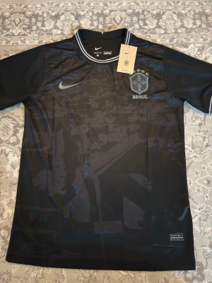 Svart Brasilien fotbollströja Nike - Snygg svart fotbollströja från Nike med Brasilien-logga och diskret mönster på framsidan. Tröjan har korta ärmar, rund hals med grå detaljer och Nike Swoosh på bröstet. Perfekt för dig som gillar sportig stil och vill visa stöd för Brasilien.