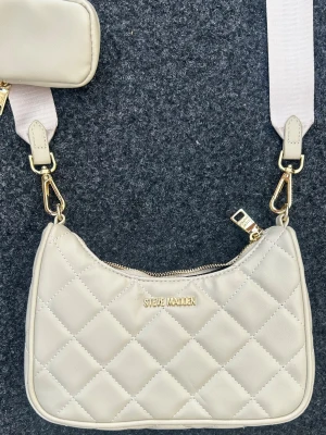 Beige quiltad axelväska Steve Madden - Snygg beige axelväska från Steve Madden med quiltat mönster och guldfärgade detaljer. Väskan har en bred axelrem och en liten avtagbar ficka på remmen. Tillverkad i konstläder och stängs med dragkedja. Perfekt för dig som gillar trendiga accessoarer.