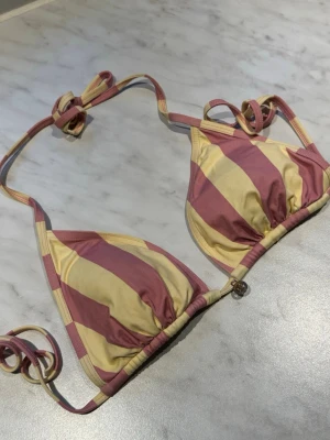 Randig bikiniöverdel i rosa och gul - Snygg trekantsbikini med breda ränder i ljusgul och rosa. Klassisk modell med knyt i nacke och rygg, samt små detaljer vid snörena. Perfekt för stranden eller poolen!