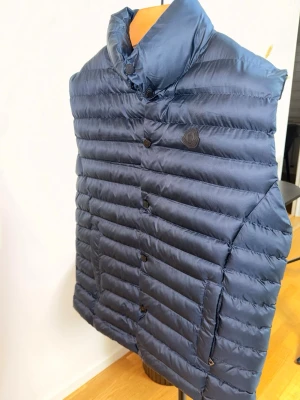 Mörkblå dunväst från Moncler - Snygg mörkblå dunväst från Moncler med hög krage och knappstängning. Västen har Moncler-logga på bröstet och två sidofickor. Tillverkad i glansigt material med horisontella sömmar för en modern look.