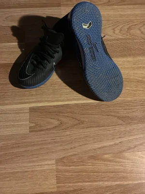 Nike svart/blå fotbollsskor Zoom - Säljer ett par Nike fotbollsskor med svart ovandel och blå sula. Skorna har snörning och Nike-logga på sidan samt Zoom-teknologi i sulan. Perfekta för futsal eller spel på hårda underlag. Materialet är syntet och gummi. 