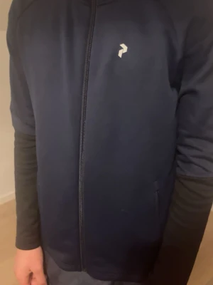 Mörkblå hoodie från Peak Performance - Snygg mörkblå hoodie från Peak Performance med dragkedja framtill och diskret logga på bröstet. Huva med textdetalj runt kanten. Tillverkad i mjukt och stretchigt material som känns skönt mot huden. Perfekt för en sportig och avslappnad look.
