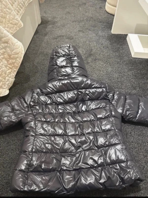 Svart pufferjacka med huva från 157 - Säljer en svart, glansig pufferjacka från 157 med huva och dragkedja. Jackan har breda, vadderade paneler och en klassisk pufferlook som håller dig varm under kalla dagar. Materialet är syntetiskt och jackan har en modern, avslappnad passform.