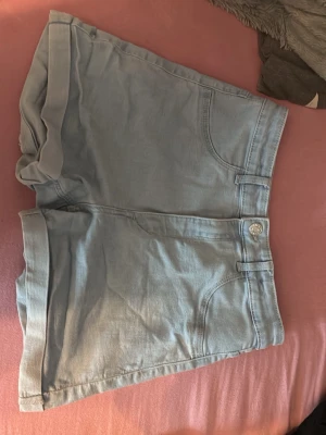 Ljusblå denimshorts med uppvik - Säljer ett par ljusblå denimshorts med uppvikta benslut och klassisk femficksdesign. Shortsen har knapp och dragkedja framtill och är perfekta för varma dagar. Passar till det mesta och är riktigt sköna att bära.