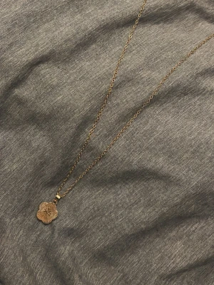Guldigt halsband med blomma - Snyggt halsband i guld med tunt kedja och ett hängsmycke i form av en blomma. Ena sidan av hänget har en räfflad yta medan den andra är slät och blank. Perfekt accessoar för dig som gillar stilrena och feminina detaljer.