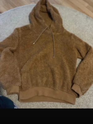 Brun fluffig hoodie med huva - Mysig brun hoodie i fluffigt material med huva och dragsko. Tröjan har ribbade muddar vid ärmslut och nederkant samt en stor magficka. Perfekt för dig som gillar en avslappnad och trendig stil.