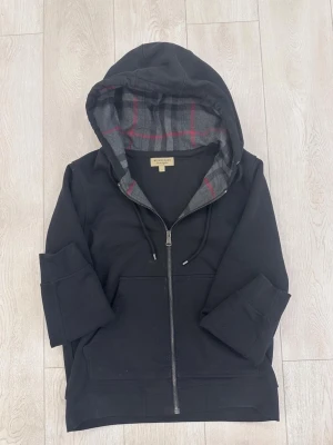 Svart hoodie från Burberry med dragkedja - Snygg svart hoodie från Burberry med dragkedja framtill och klassisk rutmönstrad insida i huvan i grått och rött. Hoodien har ficka framtill och justerbar dragsko i huvan. Perfekt för en avslappnad och stilren look.