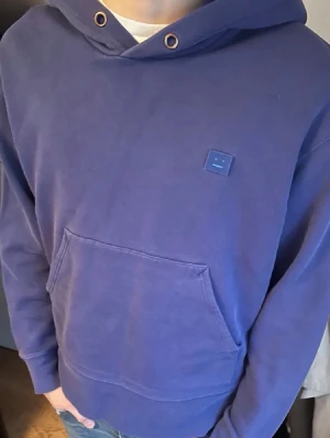 Blå hoodie från Acne Studios - Säljer en blå hoodie från Acne Studios med klassisk huva och känguruficka. På bröstet sitter Acne Studios ikoniska ansiktslogga i matchande blått. Hoodien har ribbade muddar och är gjord i mjukt bomullsmaterial. Perfekt för en chill och stilren look.