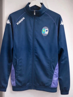 Blå träningsjacka från Kappa - Mörkblå träningsjacka från Kappa med dragkedja, ståkrage och klubbmärke på bröstet. Jackan har vita Kappa-loggor på axlarna och lila paneler vid sidorna. Perfekt för träning eller som sportig vardagsstil.