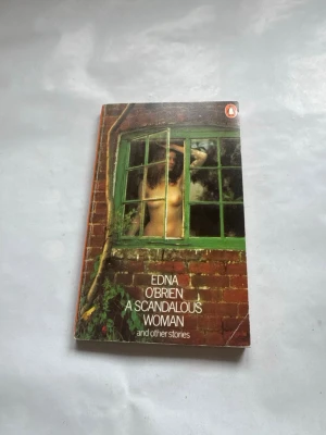 A Scandalous Woman av Edna O'Brien - En engelsk pocketbok med titeln 'A Scandalous Woman and other stories' av Edna O'Brien. Omslaget har ett konstnärligt foto av en kvinna bakom ett grönt fönster i en tegelvägg. Perfekt för dig som gillar klassisk skönlitteratur.