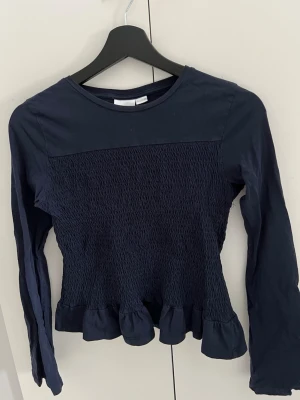 Mörkblå långärmad topp med volang från Gina Tricot - Mörkblå långärmad topp med smockad framsida och baksida samt volangkant nedtill. Rund halsringning och figurnära passform. Även fina vida armar nertill. Perfekt för dig som gillar stilrena detaljer och en feminin touch. Använd några gånger men fortfarande topp skick!