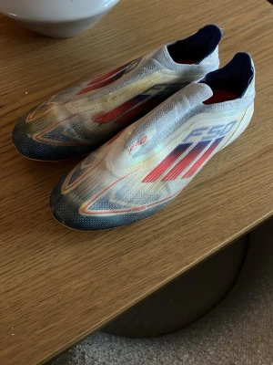 Adidas F50 elite  - Säljer ett par Adidas F50 fotbollsskor i vitt med röda och blå detaljer. Skorna har en strömlinjeformad design utan snörning och är tillverkade i ett lätt syntetmaterial för maximal fart på planen. Perfekta för dig som vill sticka ut med både stil och prestanda. Lite slitna på framsidan men inget som märks på planen 