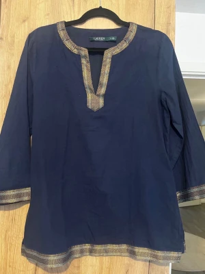 Mörkblå blus med gulddetaljer Ralph Lauren - Snygg mörkblå blus från Lauren med guldfärgade broderade detaljer vid halsringning, ärmslut och nederkant. Blusen har v-ringad öppning och trekvartsärm. Perfekt för dig som vill ha något unikt och stilrent. Stl M