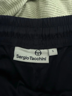 Marinblå byxor från Sergio Tacchini - Snygga marinblå shorts från Sergio Tacchini med elastisk midja och logga broderad på sidan. Lätta och bekväma, perfekta för varma dagar eller sport. Klassisk design med fickor och diskret dragkedja på sidan.