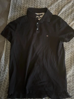 Svart pikétröja från Tommy Hilfiger - Klassisk svart pikétröja från Tommy Hilfiger med liten logga på bröstet och knappar vid halsen. Tröjan har korta ärmar och mjukt bomullsmaterial som känns skönt mot huden. Perfekt till jeans eller shorts för en clean look.