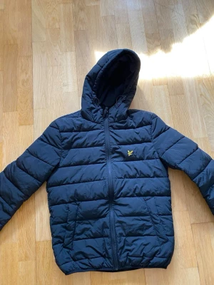 Mörkblå pufferjacka från Lyle & Scott - Snygg mörkblå pufferjacka från Lyle & Scott med gul logga på bröstet. Jackan har huva, dragkedja framtill och två sidofickor. Quiltad design med breda kanaler och elastiska muddar vid ärmslut och nederkant. Perfekt för kalla vinterdagar.