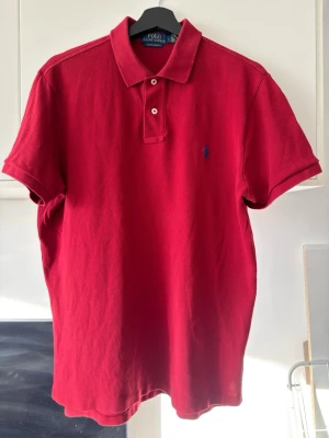 Röd pikétröja från Polo Ralph Lauren - Klassisk röd pikétröja från Polo Ralph Lauren med korta ärmar och blå broderad logga på bröstet. Tröjan har krage och två knappar framtill. Tillverkad i bomull för en bekväm och stilren look.