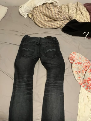 Svarta slim fit jeans från Nudie jeans - Säljer ett par svarta slim fit jeans från Nudie Jeans med klassiska fem fickor och snygga slitningar. Jeansen har Nudies ikoniska broderi på bakfickorna och är tillverkade i stretchigt denimtyg för skön passform.