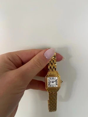 Guldfärgad klassisk klocka - Snygg guldfärgad klocka med klassisk länkarmband och fyrkantig urtavla. Urtavlan är vit med svarta romerska siffror och blå visare. Elegant och tidlös design som verkligen sticker ut. Klockan är aldrig använd. Extra länkar finns. Batteriet är fullt. 