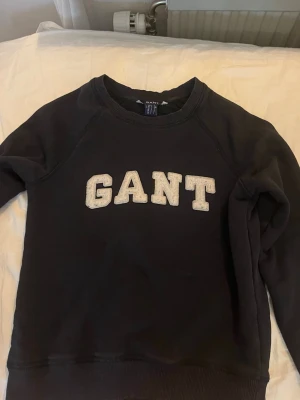 Svart/marinblå GANT hoodie med logga - Säljer en svart hoodie från GANT med stor vit broderad logga på bröstet. Klassisk modell med ribbade muddar vid ärmslut och nederkant. Perfekt för dig som gillar stilrena och tidlösa plagg med collegekänsla.