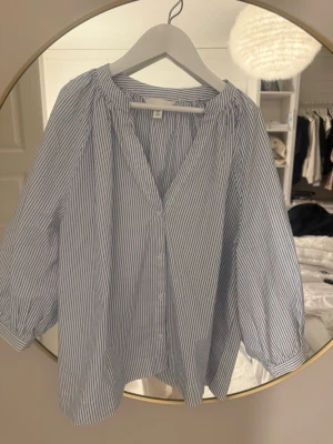 Randig blus från H&M med knappar - Säljer en luftig blus från H&M i vitt med tunna blå ränder. Blusen har v-ringning, knäppning framtill och trekvartsärmar med resår vid ärmslut. Perfekt till jeans eller kjol för en fräsch och enkel look.