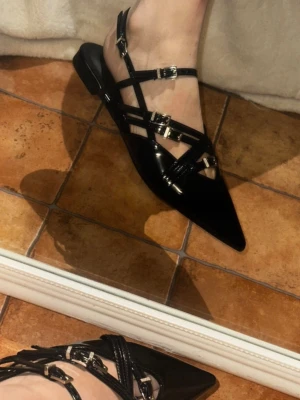 Svarta spetsiga ballerinaskor med remmar - Säljer ett par svarta ballerinaskor i glansigt material med spetsig tå och flera tunna remmar med silvriga spännen. Skorna har öppen häl och låg klack, vilket ger en edgy och trendig look. Perfekta för dig som vill sticka ut med detaljerade skor.