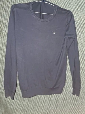 Mörkblå rundhalsad tröja från Gant - Klassisk mörkblå tröja från Gant med rund halsringning och diskret logga broderad på bröstet. Tröjan har långa ärmar och ribbade muddar vid ärmslut och nederkant. Perfekt för en stilren och enkel look. Är öppen för prisförslag vid snabb försäljning. 