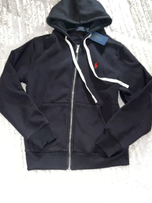 Svart hoodie från Polo Ralph Lauren - Svart hoodie med dragkedja från Polo Ralph Lauren. Klassisk design med vit dragsko i huvan, röd broderad logga på bröstet och mjukt fleecefoder på insidan. Två fickor framtill och ribbade muddar vid ärmar och nederkant. Perfekt för en avslappnad stil. Passar s